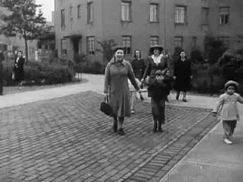 Children walking with adults on a paved courtyard outside apartment blocks
