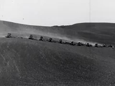 Tractors plowing a large rolling field, kicking up dust