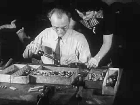 Man assembling small parts at a workbench with another person assisting