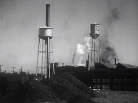 Industrial area with a water tower, smokestack, and smoke in the background.