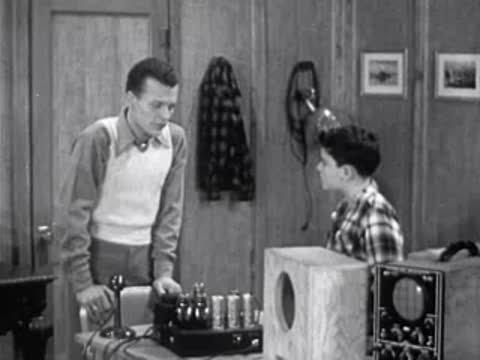 Man and boy in an amateur radio room talking near electronic equipment.