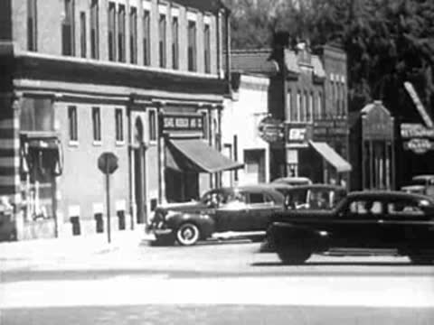 Vintage cars driving through a sunny town street intersection in the 1940s.