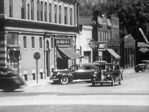 Vintage automobiles moving through a busy downtown intersection on a sunny day.