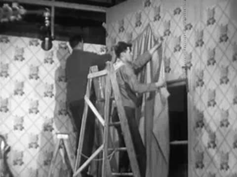 Two men on ladder hanging curtains in a set or room interior