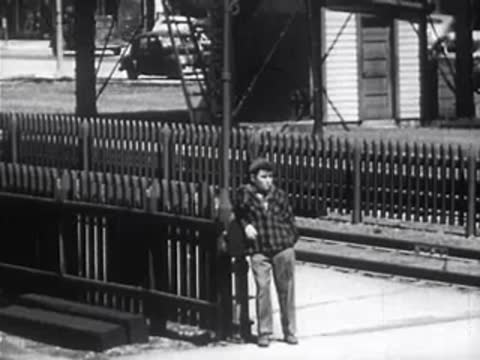 Boy in plaid shirt leaning against a fence near railroad tracks.