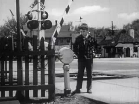 Boy standing by a railroad crossing gate and a white container on a post.