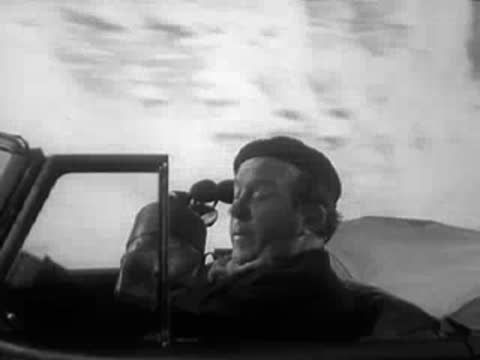 Driver with binoculars looking ahead from a convertible car.