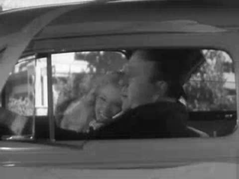 A couple sits in a vintage car as it pulls away from the intersection.