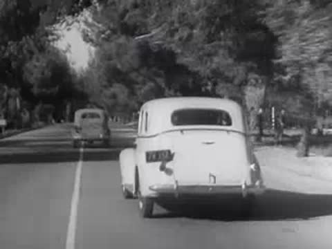A vintage car is driven down a sunlit suburban street.