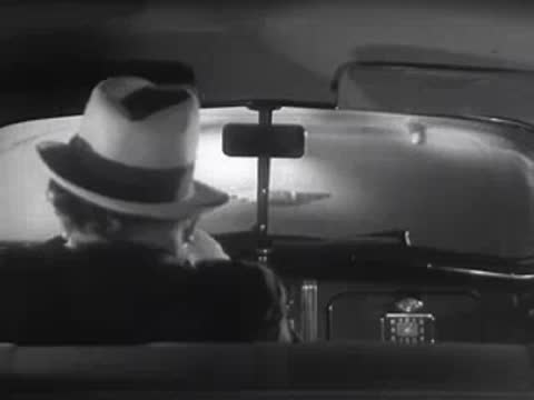 A driver in a hat looks through the rear window of a car.