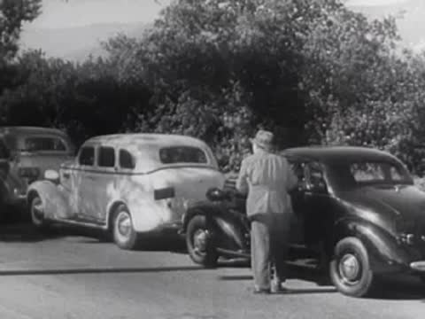 A man steps out of a parked vintage car onto the street.
