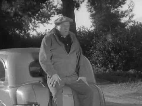 A person in work clothes sits on the rear of a car next to another car.