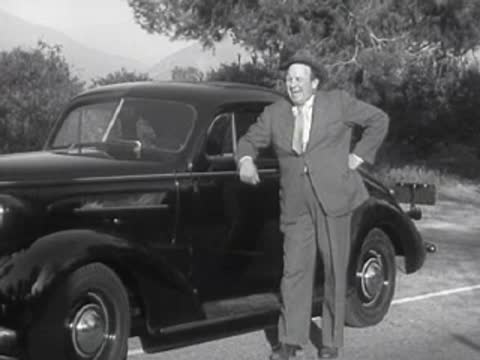 A driver looks forward inside a car while another person leans on an exterior car.