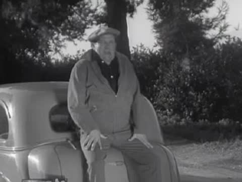 A person sits on the back of a car while a driver looks out from inside.