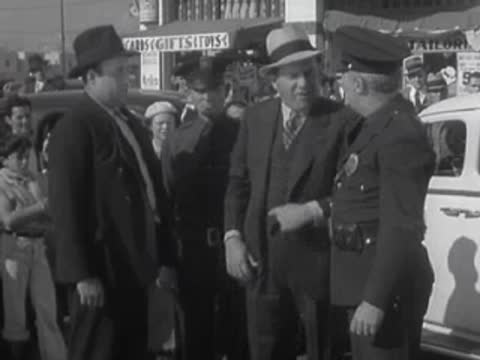 Several men stand on a city street arguing near parked cars and police officers.