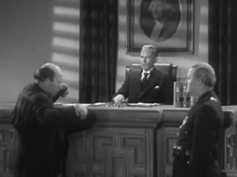 Two men converse across an office desk while a third person looks on.