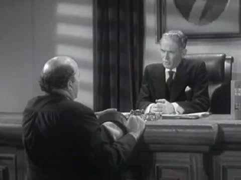 A man in a coat sits across a desk talking to an official in an office.