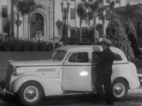 A white sedan is parked on a street lined with trees and a building entrance.