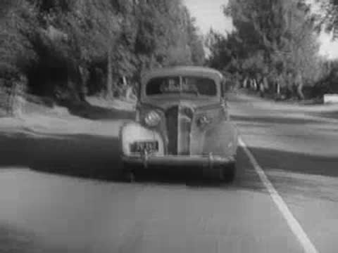 Vintage car approaching camera on a paved tree-lined road.