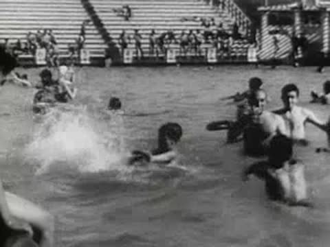 people swimming and diving in an outdoor pool with spectator stands