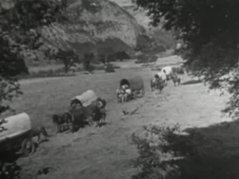 horse-drawn covered wagons moving along a dirt path through a grassy valley