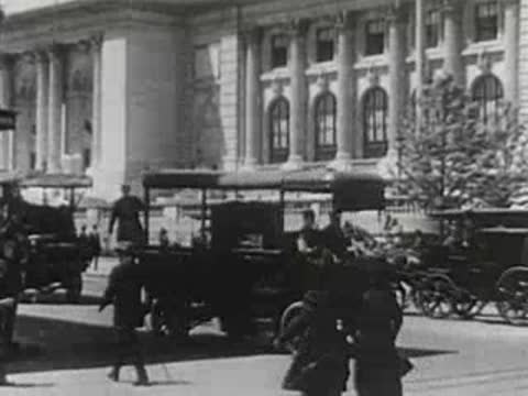 Vintage car driving past horse-drawn carriages and pedestrians on a city street