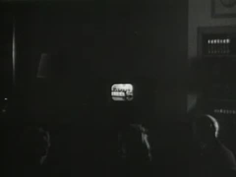 Darkened room with audience watching a small illuminated television screen, early television broadcast viewing