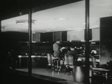 Control room with personnel working at consoles behind large glass windows, 1940s.
