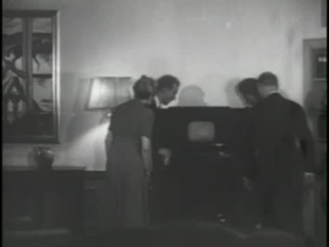People gathered around an early television console in a living room.