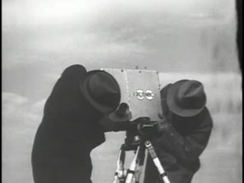 Two men in hats set up a television camera on a tripod outdoors.