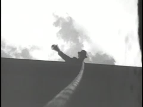 Silhouette of a man reaching up along a cable against a cloudy sky.