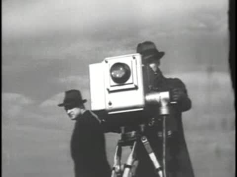 A television camera on a tripod outdoors with operators nearby.