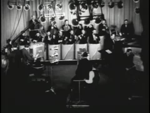 Orchestra performing on stage with studio lights and crew in background