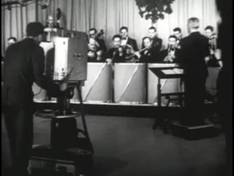 Conductor leading an orchestra on a studio stage with camera in foreground
