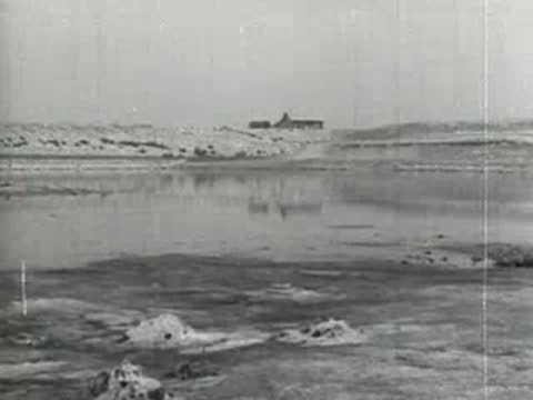 Still body of water reflecting dry landscape and distant small building.