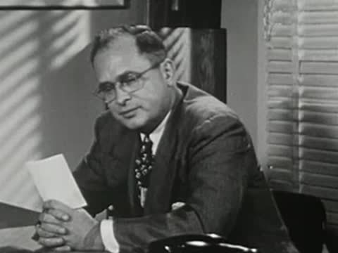 Man in suit at office desk talking while holding papers.
