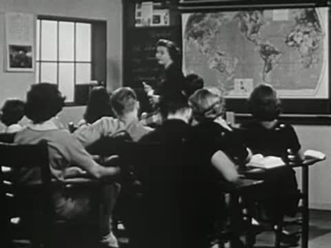 Teacher standing before world map pointing in crowded classroom.