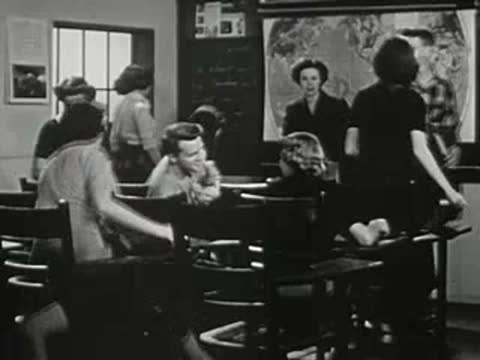 Students standing and moving around desks in crowded classroom.