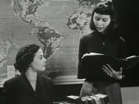 Two female students in front of world map looking at book.
