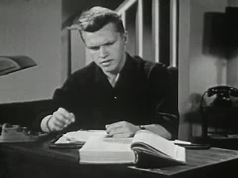 Young man sitting at desk writing in large book.