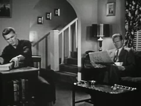 Man reading newspaper on couch as teenager gets up from desk in background.