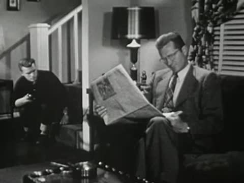 Man reading newspaper on couch while teenager talks on phone nearby.