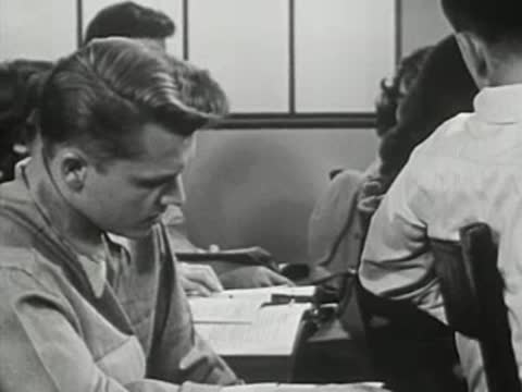 Student working at desk in classroom with other students around him.