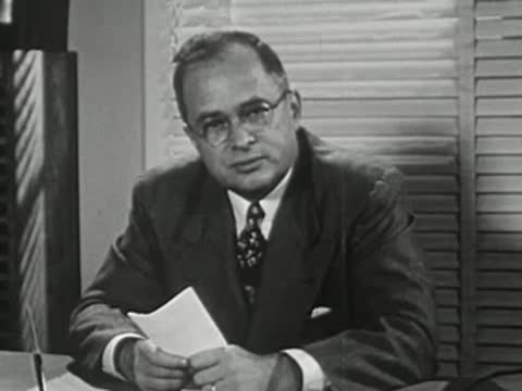 Adult man with glasses sitting at desk holding papers and speaking.