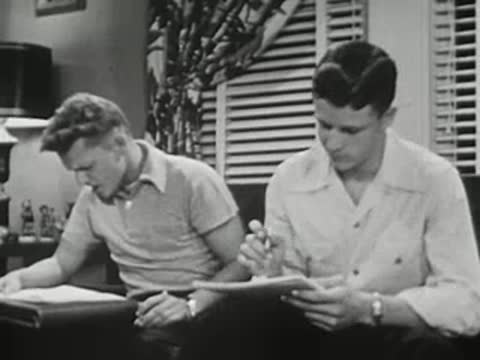 Two young men studying together at table, writing in notebooks.