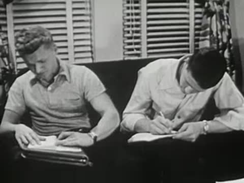 Two young men sitting on couch studying and writing notes intently.