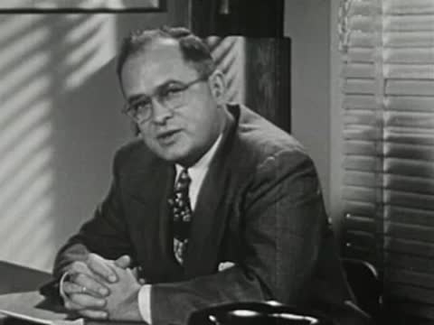 Adult man in suit with glasses sitting at desk with folded hands.