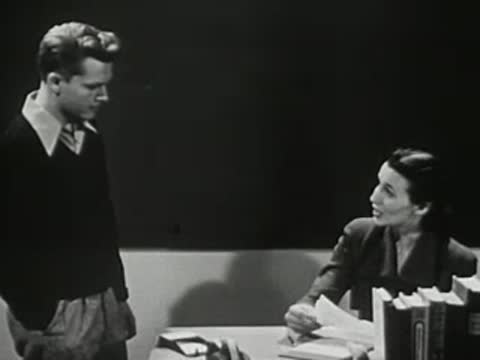 A male student talking to a female teacher sitting at her desk