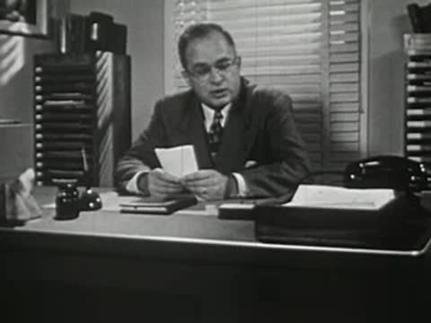 Man in suit sitting at office desk talking while holding papers.