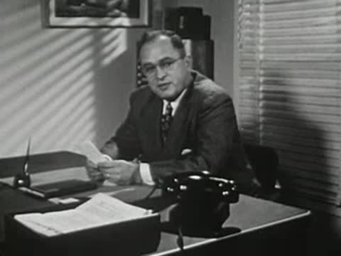 Man in suit at office desk talking next to telephone and lamp.